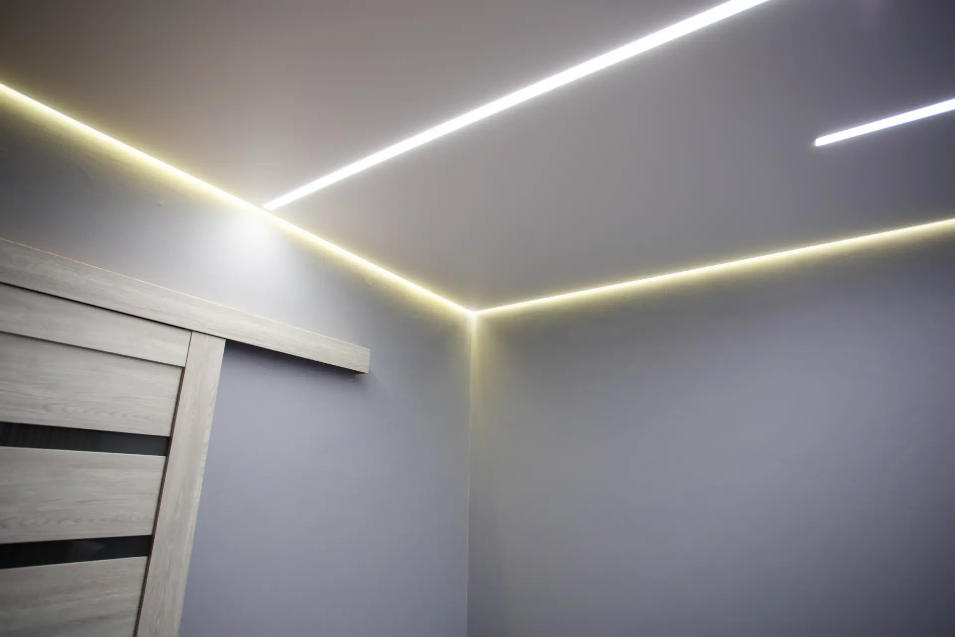 Moderný napínaný strop s integrovaným LED pásovým osvetlením v rohoch miestnosti, vytvárajúci jemné ambientné svetlo a elegantný dizajn interiéru.