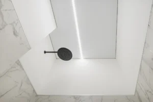 Minimalistický biely strop v sprche s čiernou hlavovou sprchou a úzkym LED pásom uprostred stropu.