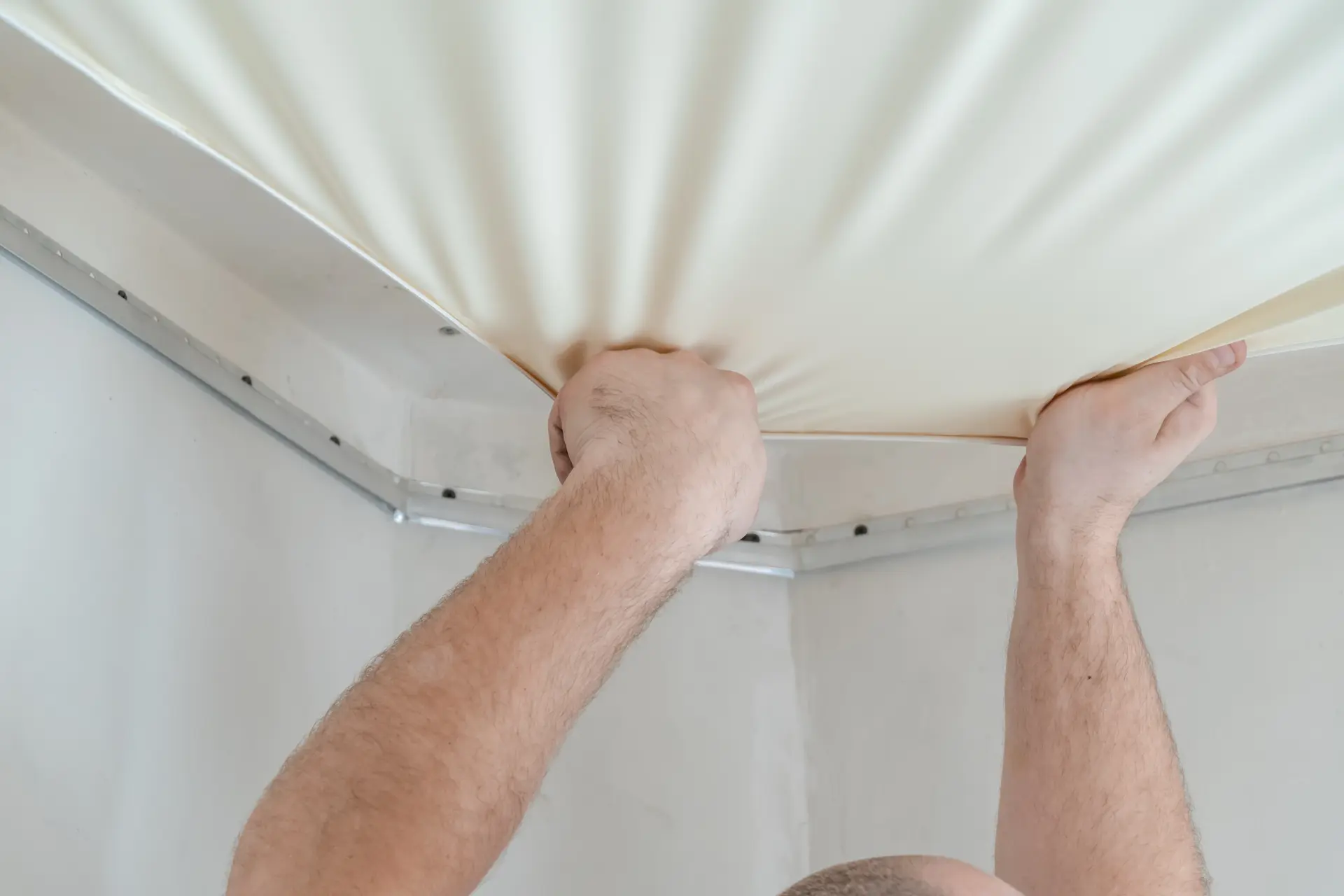 Montáž napínaného stropu – pracovník napína PVC fóliu do montážneho profilu pri strope.